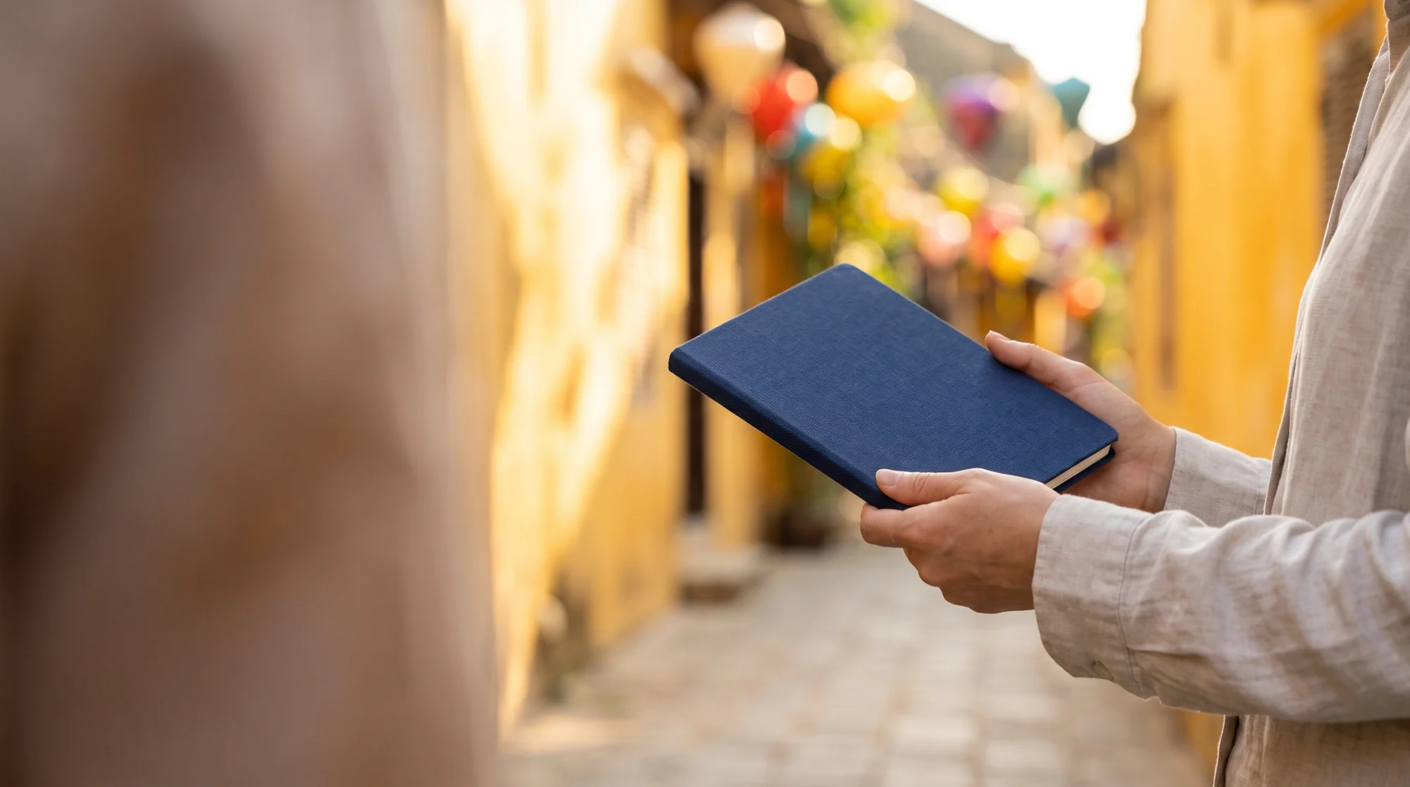 Close view of hands holding a closed navy travel booklet with Hoi An Ancient Town lanterns softly blurred behind